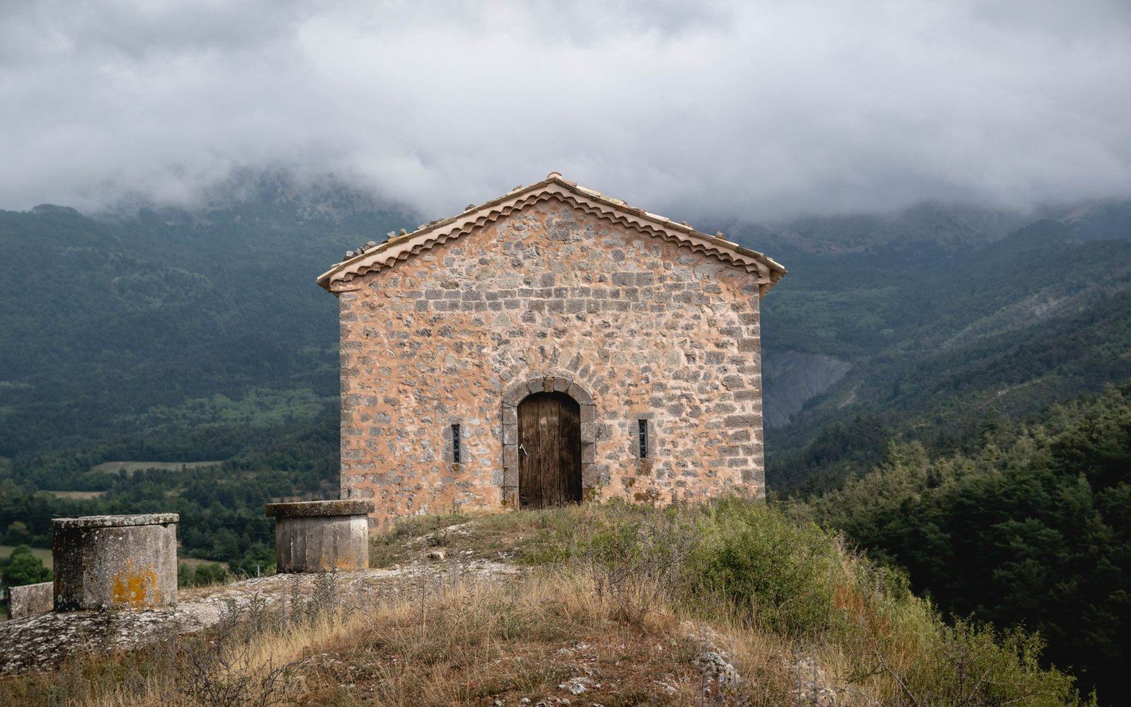 Stone chapel with tiled roof stands atop a hilltop overlooking misty forested mountains