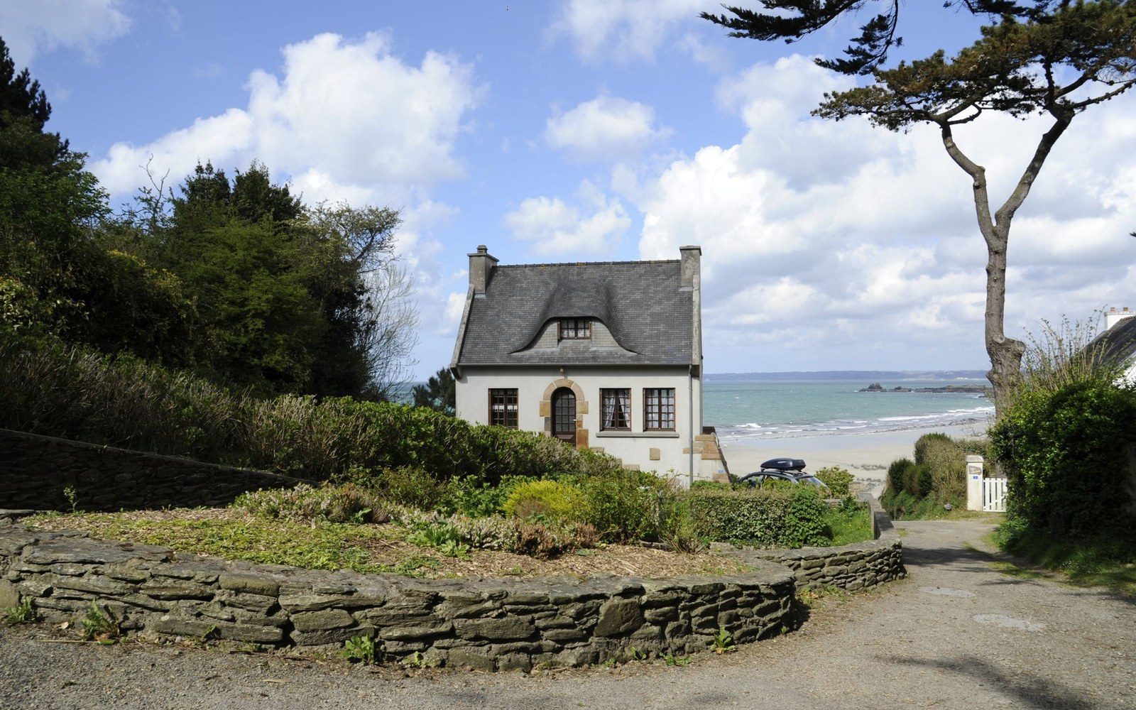Small slate-roofed cottage beside a stone wall overlooking a sandy beach and sea