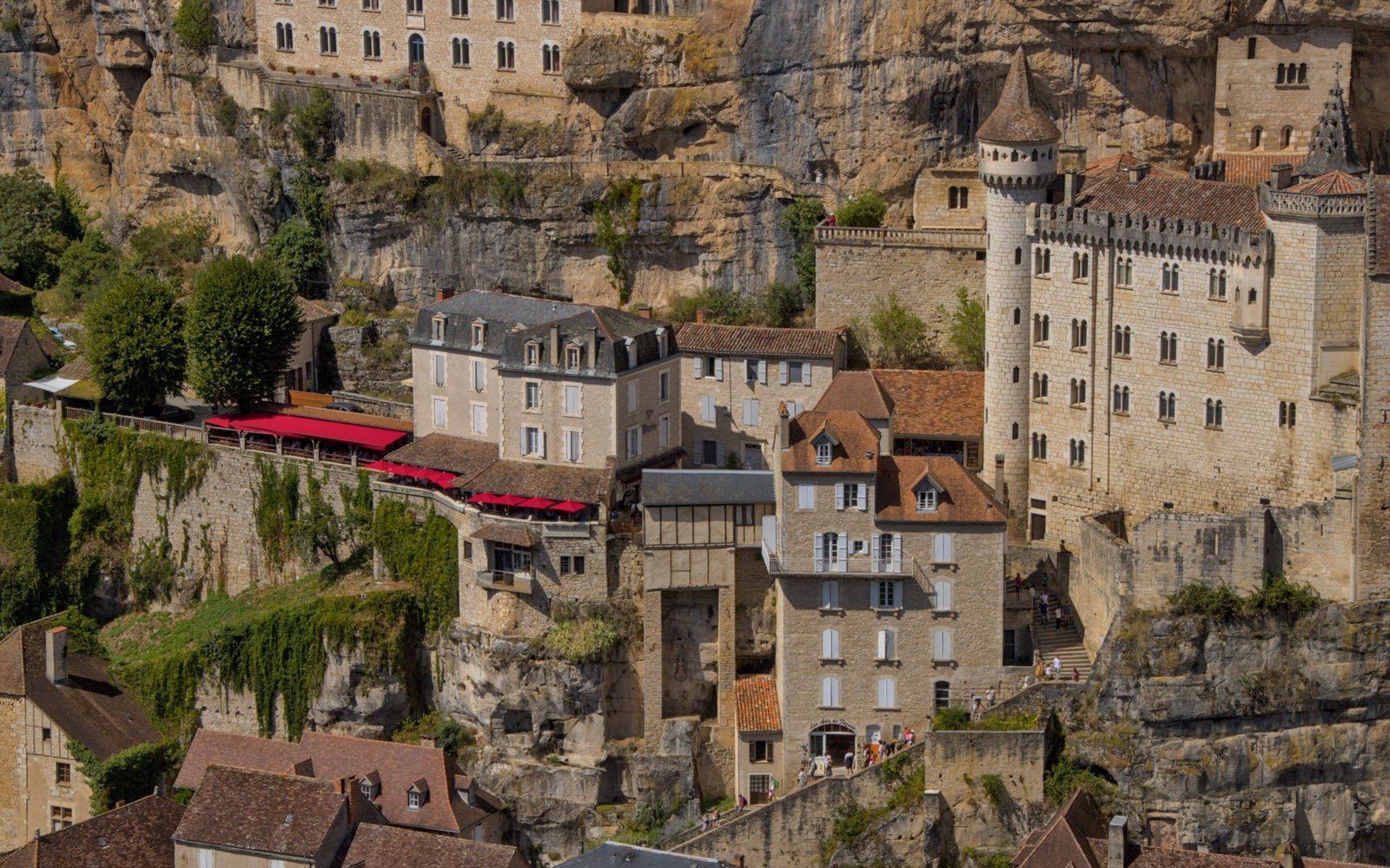 Stone buildings with a turreted château cling to a limestone cliff above a hillside village