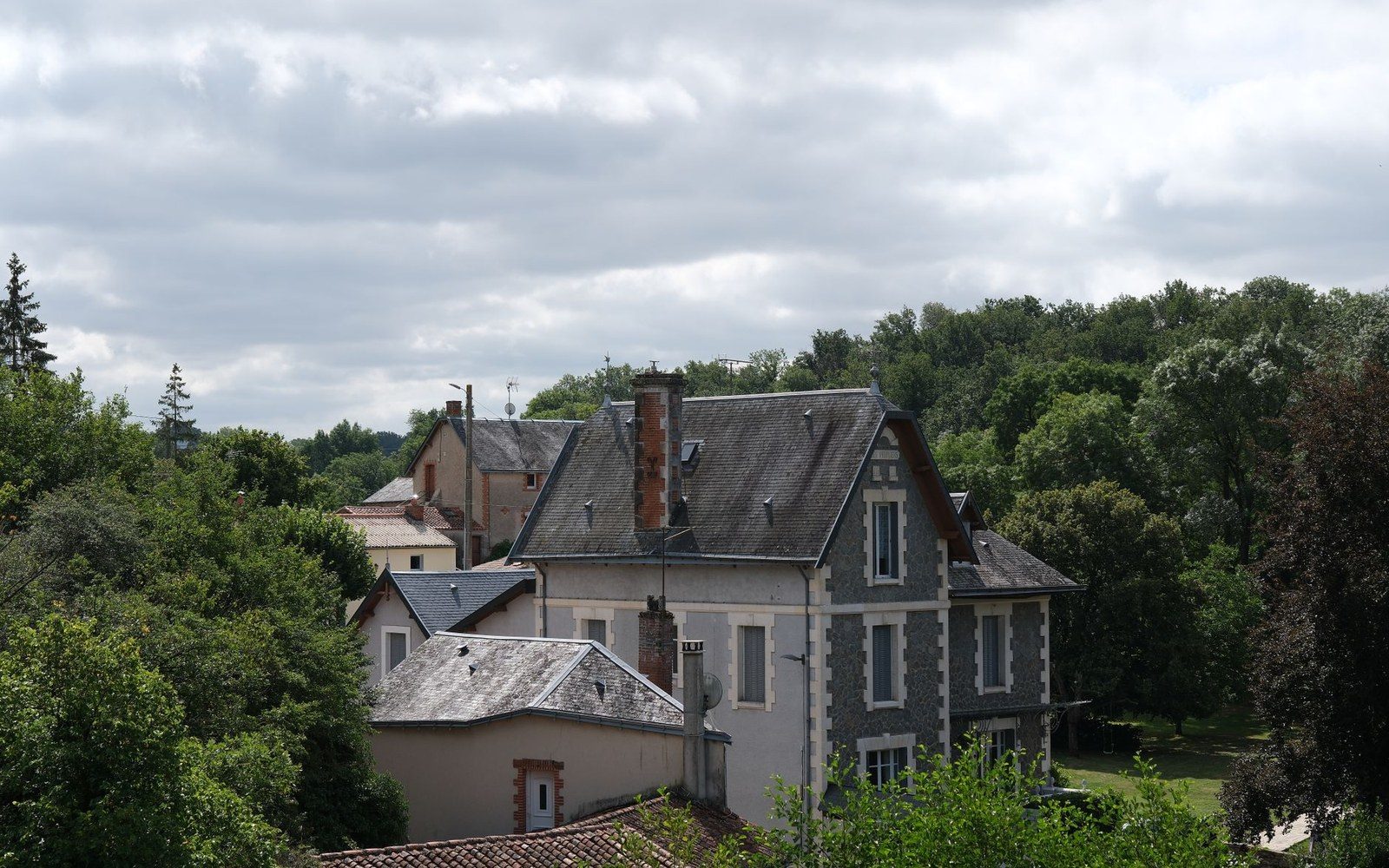 A stone house with a steep slate roof surrounded by trees under a cloudy sky