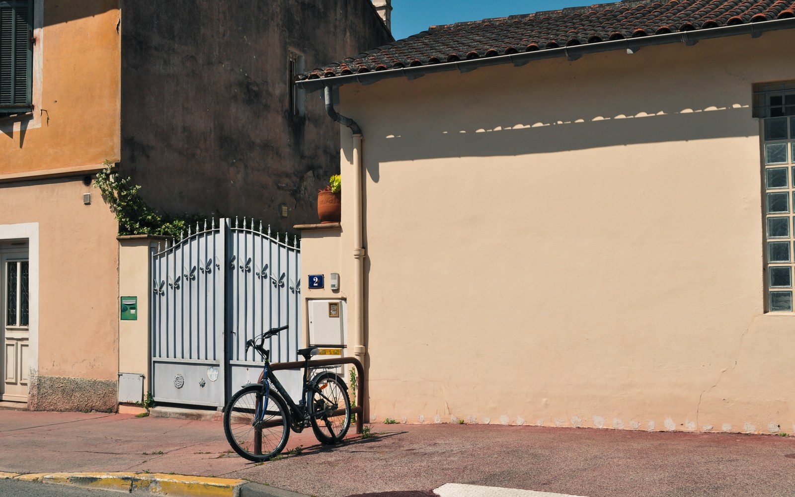 A bicycle leans against a metal gate beside a pale stucco house with terracotta roof tiles