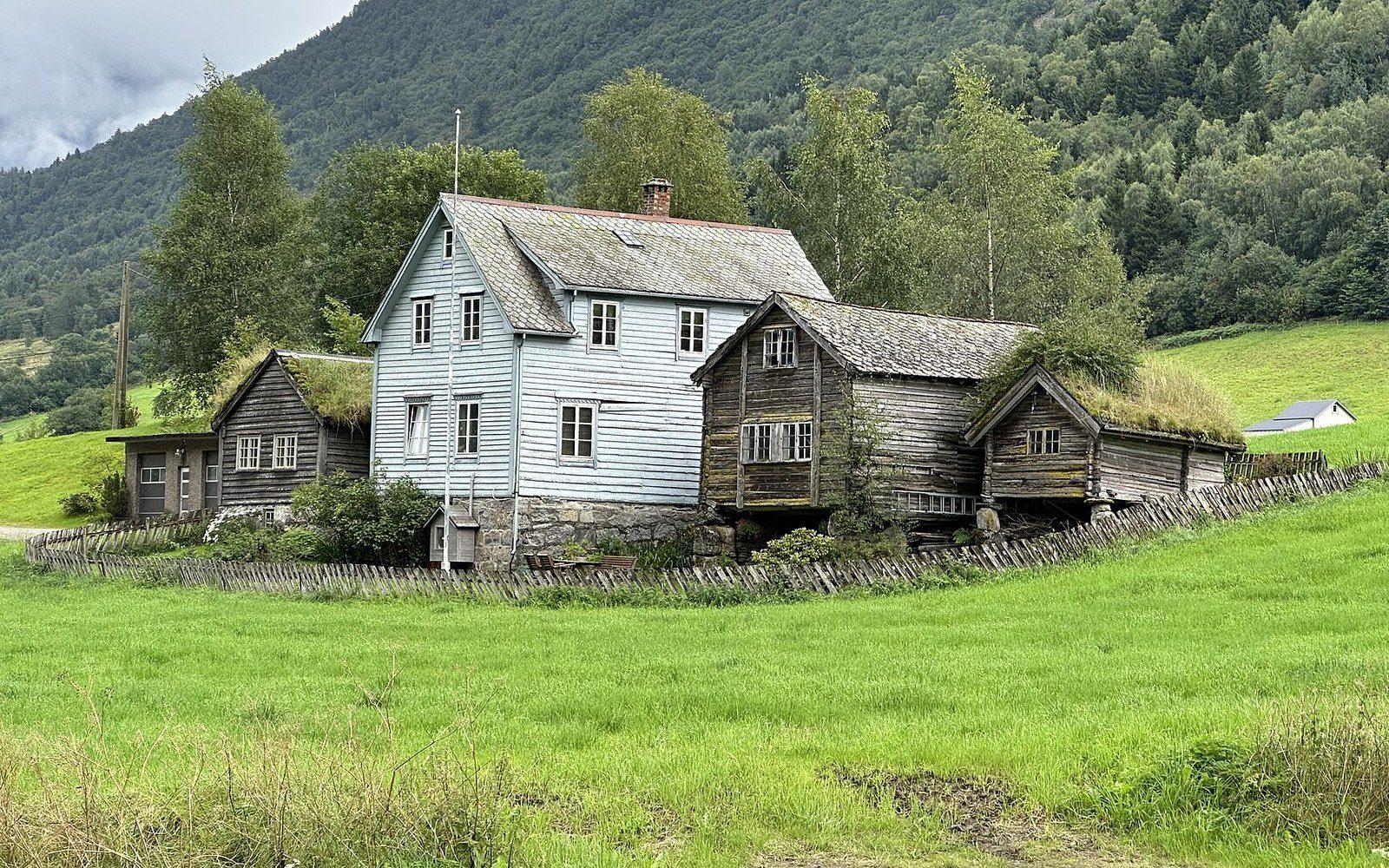 Weathered wooden farmhouse with grass-roofed outbuildings beside a green mountain field