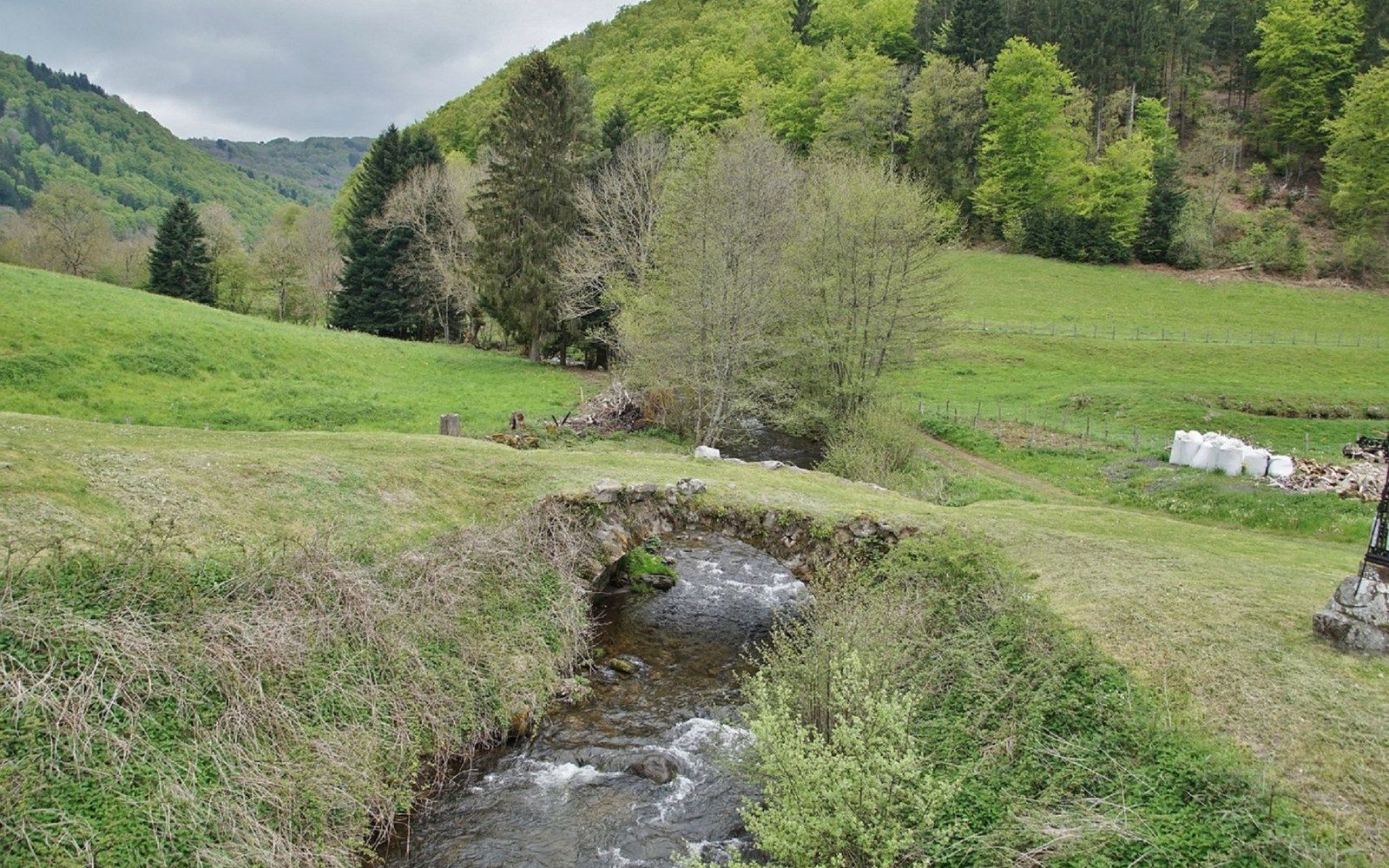 A small stone bridge arches over a stream flowing through a green valley pasture